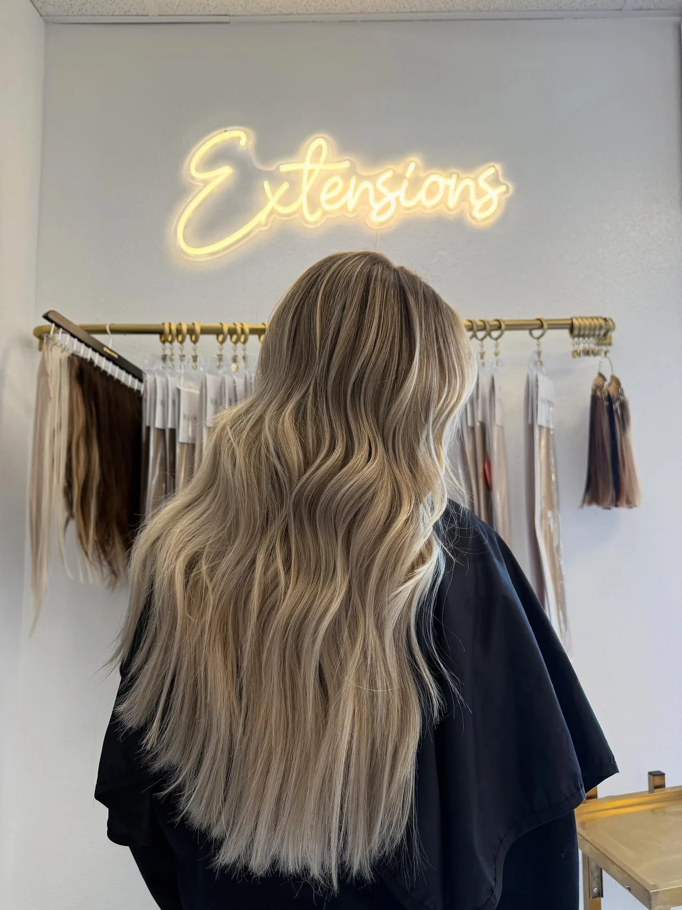 Long blonde hair with extensions blended through the mid-lengths, photographed beneath an Extensions neon sign